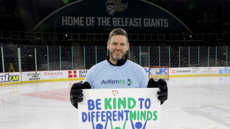 Autism NI patron stands on an ice rink holding a 'Be kind to different minds' poster