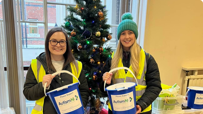 Autism NI volunteers wearing high-vis jackets and holding collection buckets in front of a Christmas tree