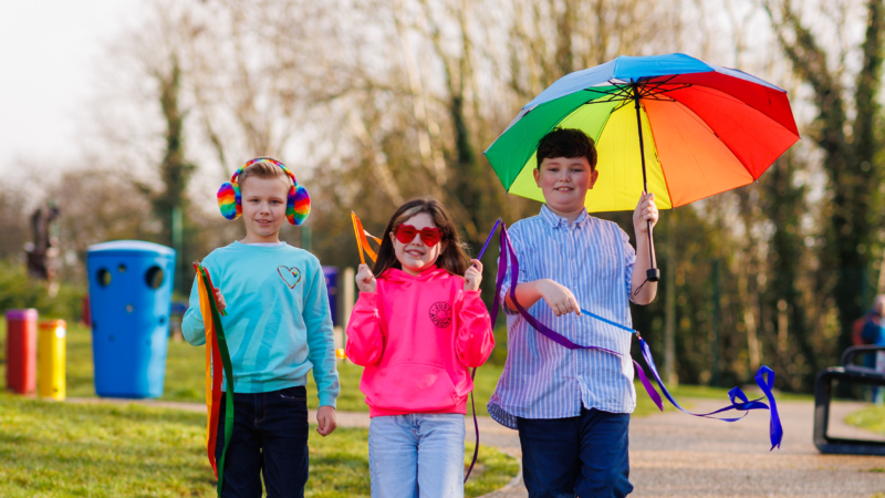 Three children in a park, wearing and holding colourful items, and walking towards the camera