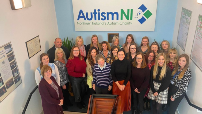 Autism NI staff members pose in a staircase in front of an Autism NI poster