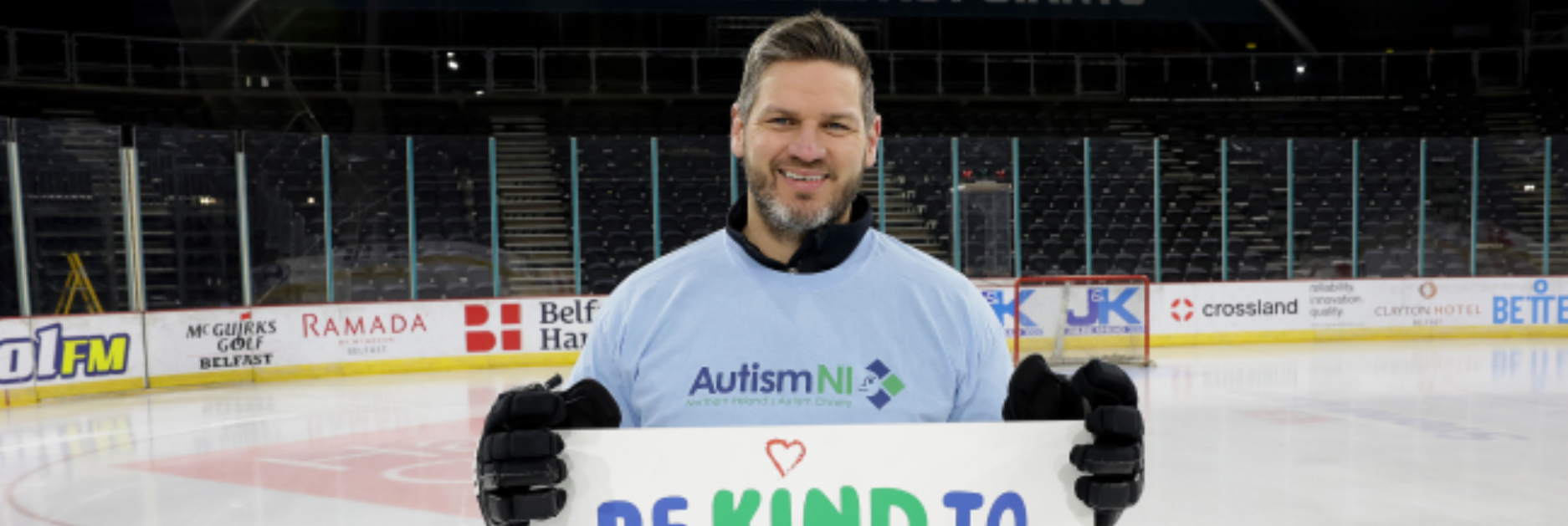 Autism NI patron stands on an ice rink holding a 'Be kind to different minds' poster