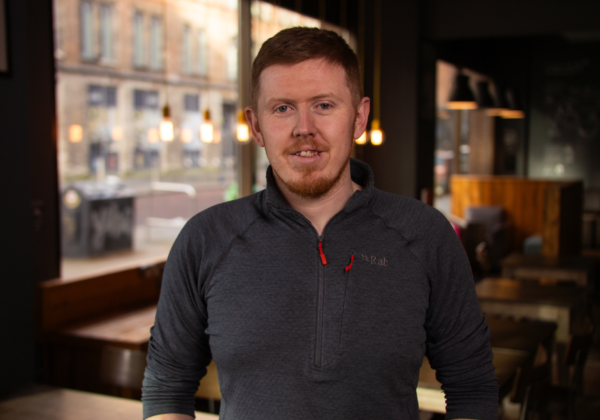 A man smiles standing in front of a café setting