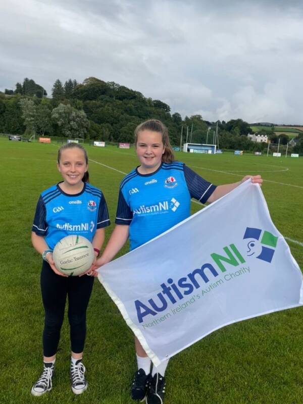 Two children standing on a pitch with a football, holding up an Autism NI flag