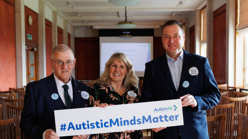 Three adults holding a sign which reads #AutisticMindsMatter