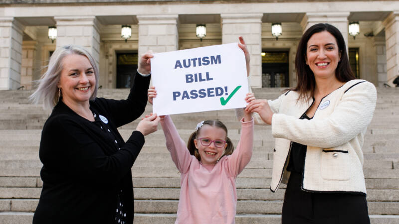 Two smiling adults and a child holding a sign which reads 'Autism Bill Passed'.