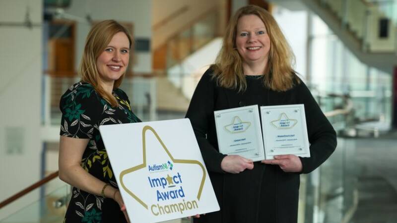Two women smile and pose with impact awards and an impact award champion poster